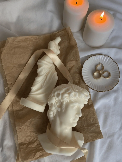 Two white sculptural candles styled on a white fabric surface with brown paper, a cream ribbon, two lit white pillar candles, a small white dish with gold rings, and a white bust-shaped candle.