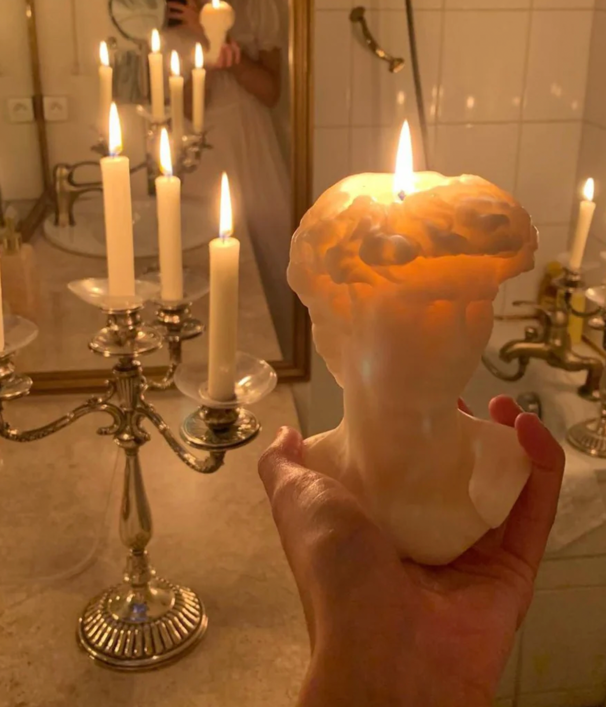 A hand holds a lit cream-colored bust-shaped candle in front of a bathroom mirror and silver candelabra.