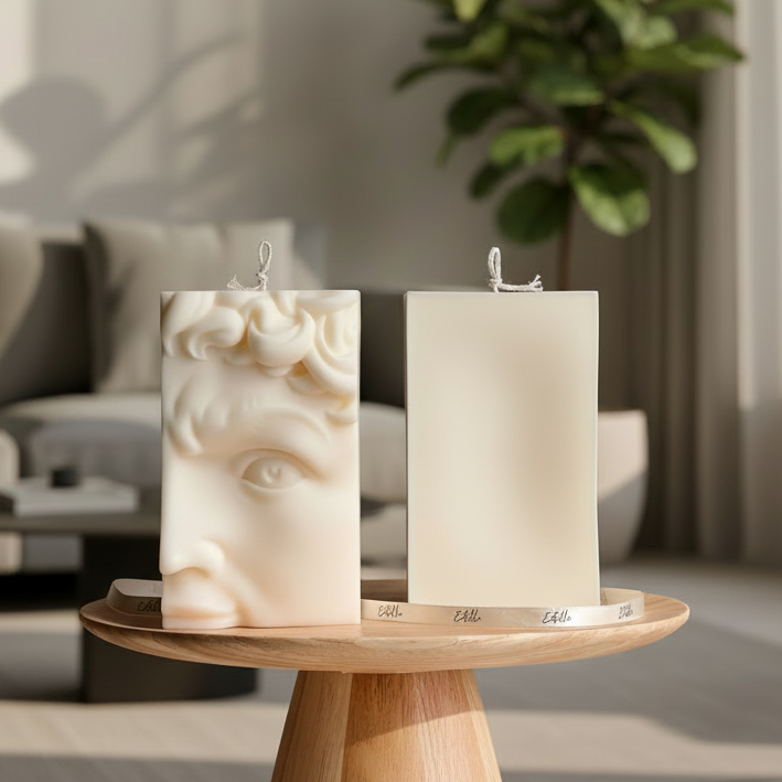 Two cream-colored candles displayed on a round wooden stand against a neutral gray background. Candle is sculpted with a classical face profile.