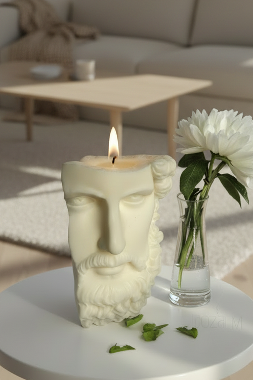 A sculptural cream-colored candle shaped like a classical male face, lit and placed on a small round white table next to a clear glass vase with white flowers in a modern, sunlit living room.