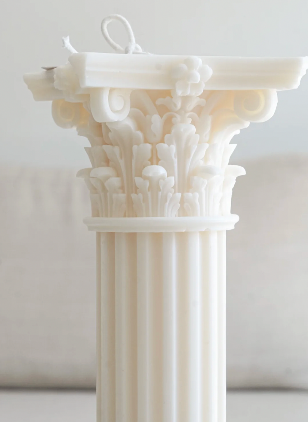 A white decorative pillar candle with an ornate classical column design, featuring a detailed capital and fluted shaft, shown against a soft, neutral background.