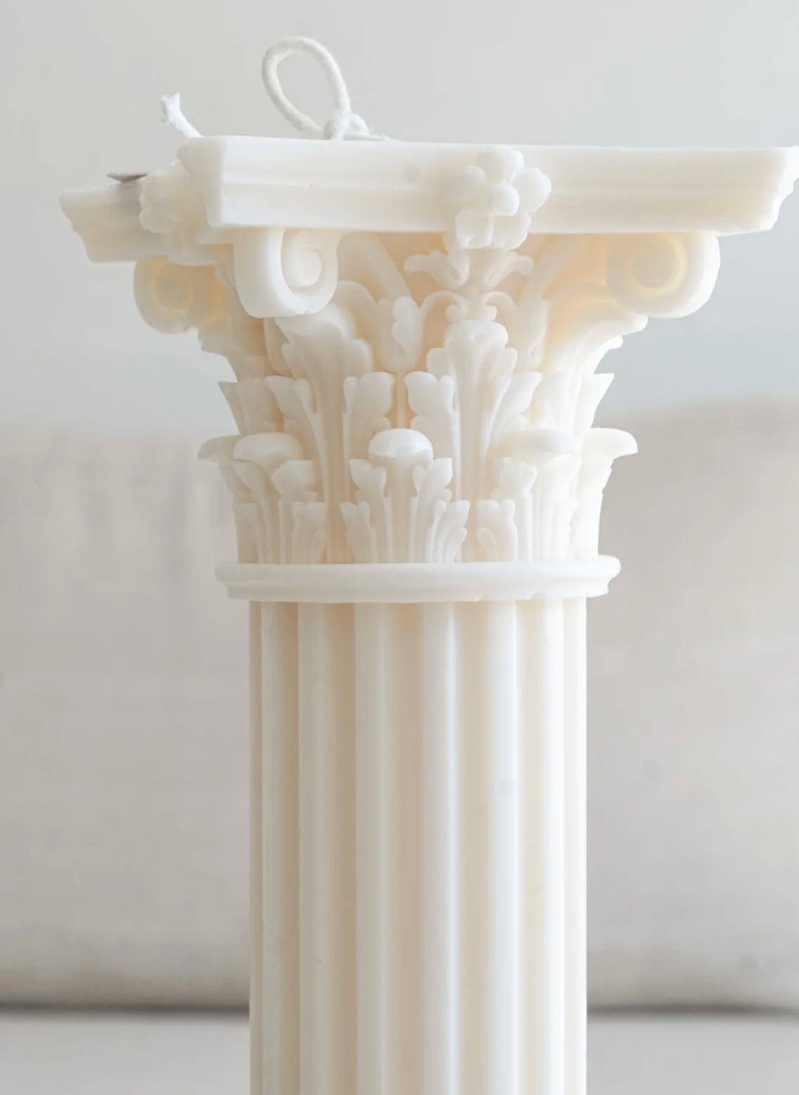 A white decorative pillar candle with an ornate classical column design, featuring a detailed capital and fluted shaft, shown against a soft, neutral background.
