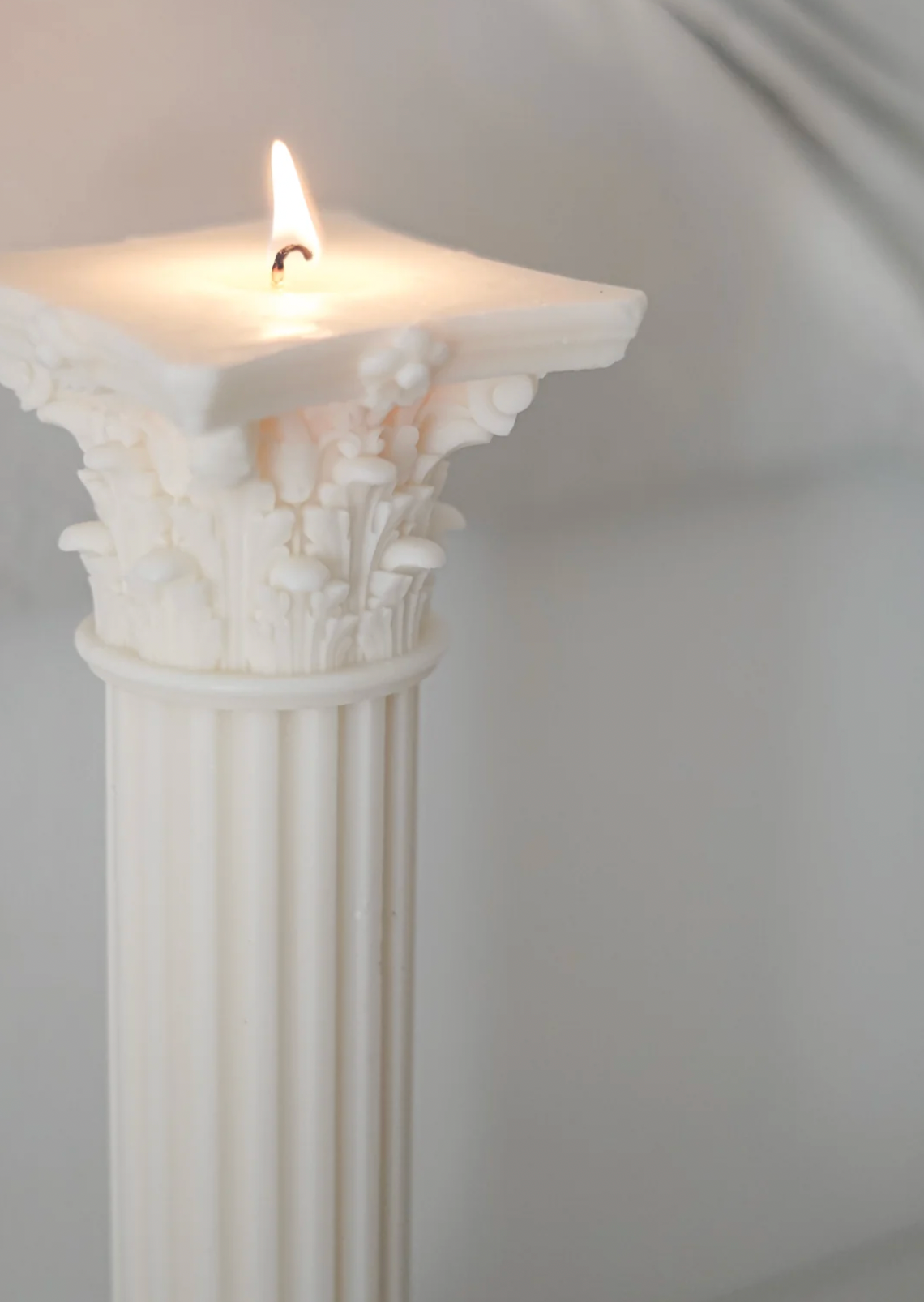 A white pillar candle shaped like a classical column with a decorative capital, shown lit with a small flame against a soft gray background.