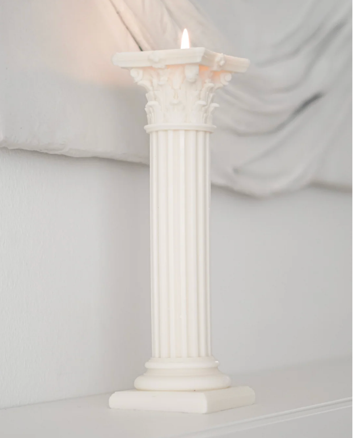 A white classical column candle holder with a lit candle, set against a soft, draped white fabric background.