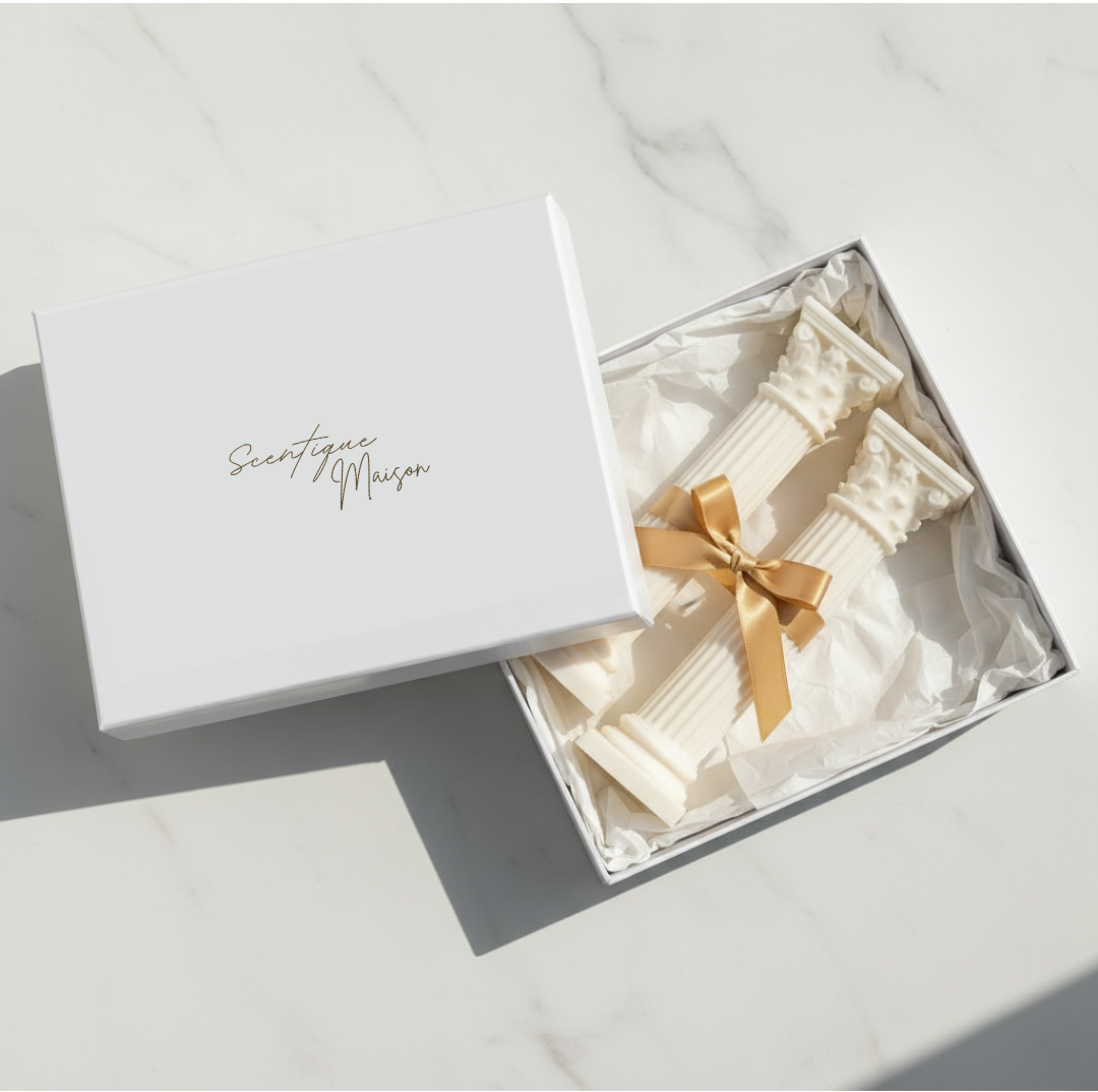 A white gift box with the brand name 'Scentique Maison in gold script, shown open on a light marble surface. Inside is a cream-colored ribbed textile gift set tied with a gold ribbon.
