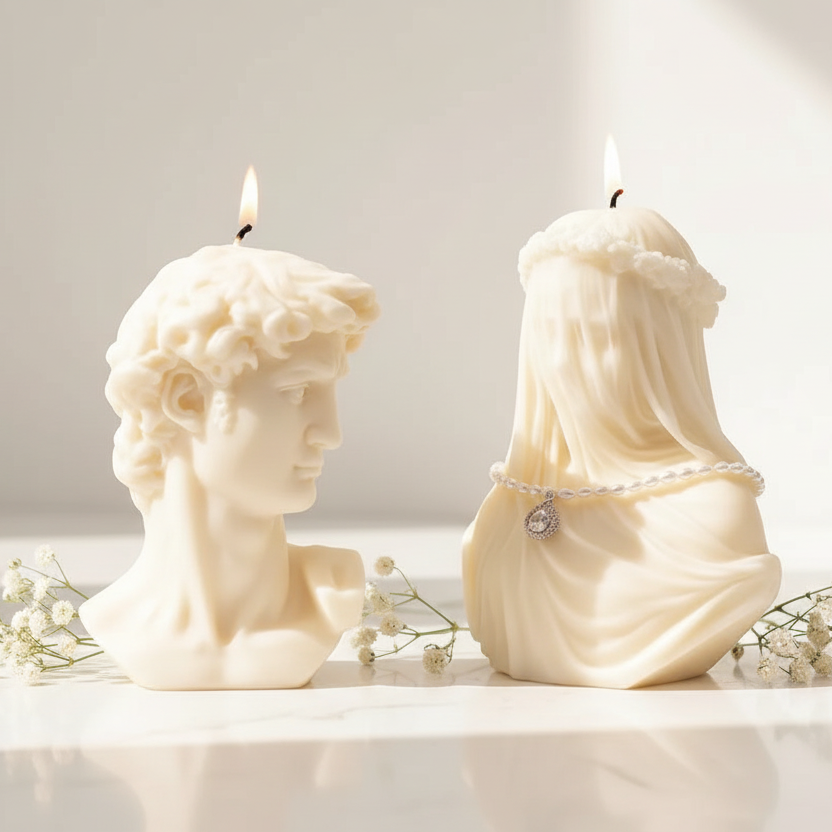 Two cream-colored sculptural candles on a white marble surface. One candle is shaped like a classical male bust, and the other is draped fabric with a veil and small pearl-like accents.
