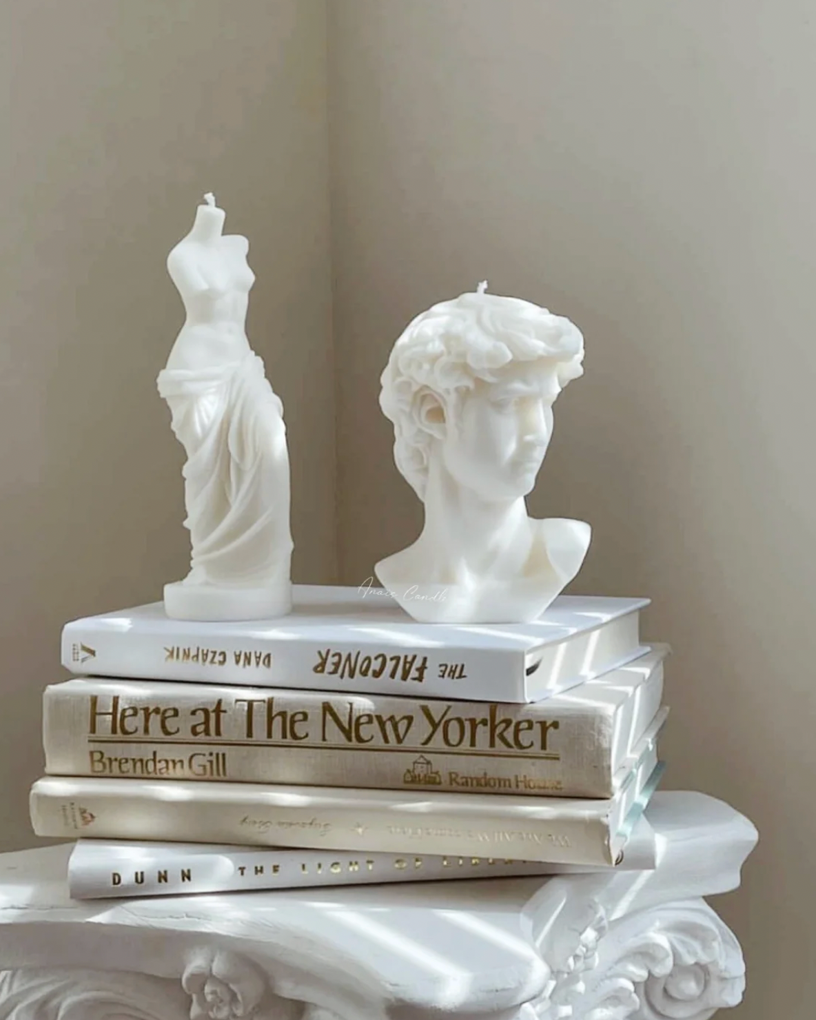 A decorative stack of white books on a white ornate surface, featuring sculptural white busts and mannequin forms on top. Visible book titles include 'The Falconer' and 'Here at The New Yorker' by Brendan Gill.